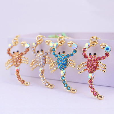 Four gold-tone scorpion charms displayed with peach, clear, blue, and pink faceted stones.