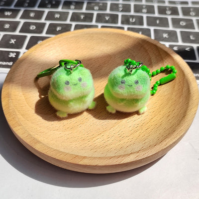 Two green flocked plush frog charms with key ring and bead chain on wood tray