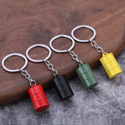 Four oil drum keychains in red, black, military green, and yellow on a wood surface.
