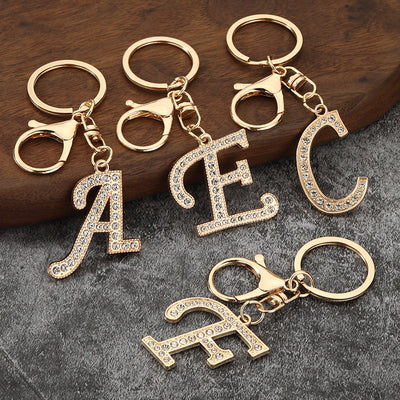 Four gold-tone clear-stone letter keychains arranged on wood and gray fabric.