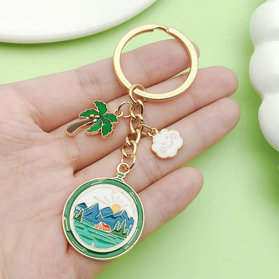 A round camping compass keychain with palm tree and cloud charms resting on a hand.