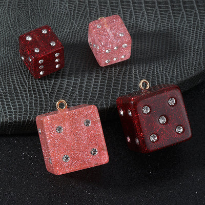 Four glitter dice charms with rhinestone dots in pink and red in two sizes on a grey and black background