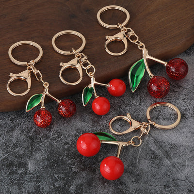 Four gold-tone cherry keychains with smooth and glitter red cherries arranged on a dark surface.
