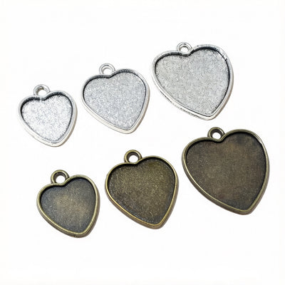 Overview of plain heart-shaped bezel trays with small top loops in silver and antique bronze tones arranged on a light b