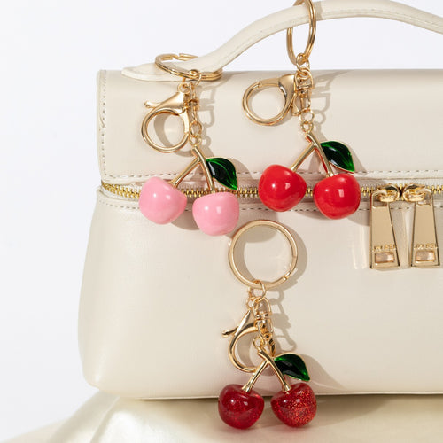 Enamel cherry keychains in pink, red and dark red clipped to a cream handbag