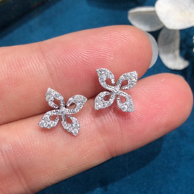 Silver four-leaf clear pave stud earrings held on fingertips over teal fabric