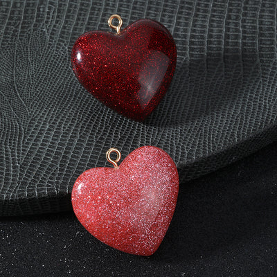 Two glitter resin heart charms, one dark red and one pink, with gold-tone jump rings placed together on a textured surfa