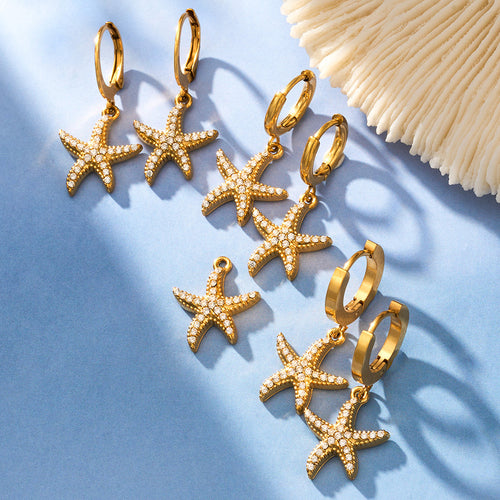 Gold starfish huggie pairs arranged on blue sand with coral