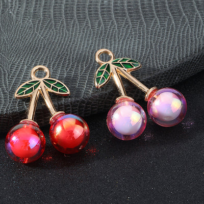 Two cherry charms with iridescent AB resin beads, one red and one pink, on a textured surface