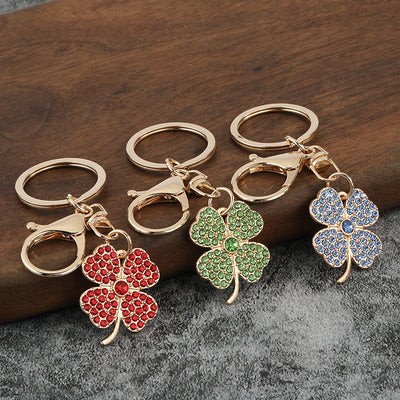 Three rhinestone clover keychains in red, green, and blue displayed on wood and gray fabric.