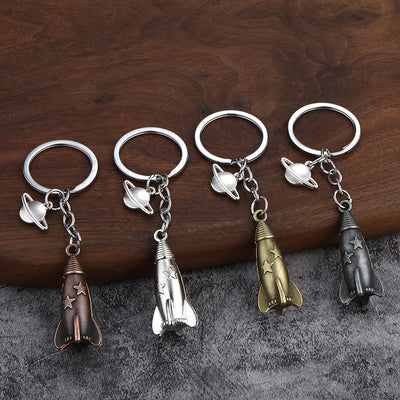 Four rocket keychains in copper, silver, bronze, and dark pewter tones displayed on wood and gray fabric.