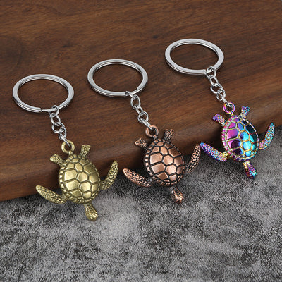 Three sea turtle keychains in antique bronze, antique copper, and rainbow finishes on wood and gray fabric.