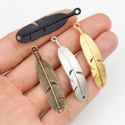 Overview of long tapered feather charms with engraved barbs in gold, silver, black and antique bronze tones arranged on 
