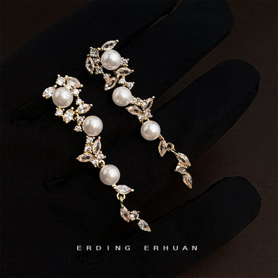 Gold-tone white pearl leaf-vine long earrings held in a black glove.