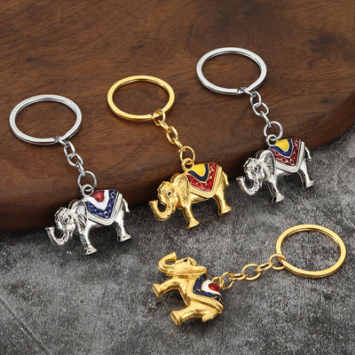 Four silver-tone and gold-tone elephant keychains arranged on wood and gray fabric.