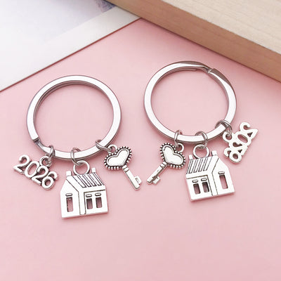 Two silver-tone keychains with house, heart-shaped key and 2026 number charms on a pink surface.