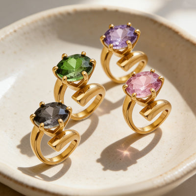 Four gold open-band rings with green black purple pink round stones on ceramic dish