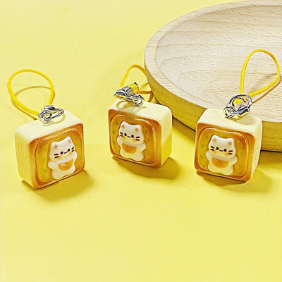 Three resin cat toast phone charms with yellow cords on yellow surface by wood dish