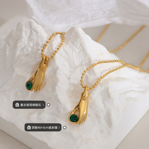 Two gold hand green zircon pendants on ball chains on white rock with labels