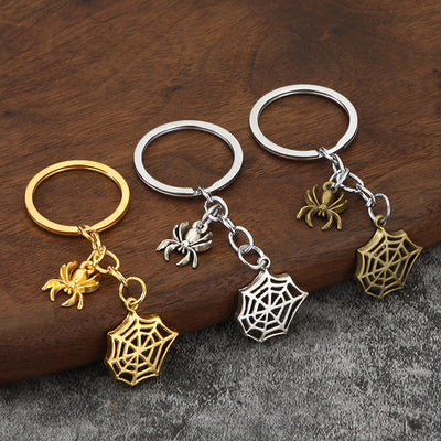 Three spider and spiderweb charm keychains in silver, antique bronze, and gold tones on wood.