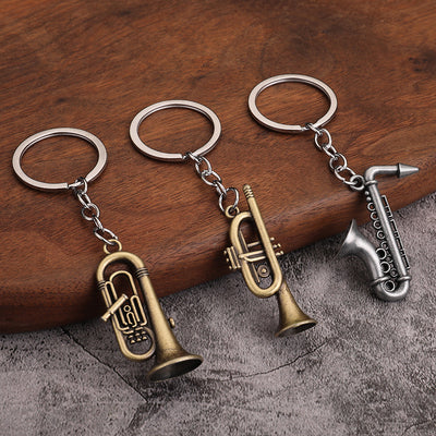 Three metal instrument keychains with rounded horn, trumpet, and saxophone charms on a wooden surface.