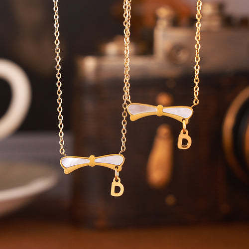 Two gold shell bow necklaces with D charms hanging against blurred camera