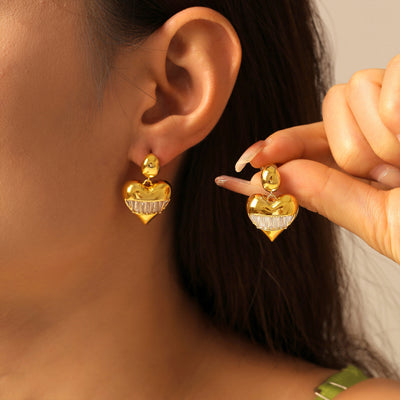 Model wearing gold heart drop earring with baguette zircon while holding matching pair