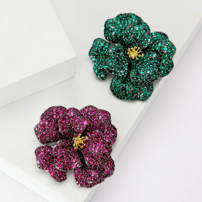 Front view of the crystal camellia brooch in purple or green with polished metal detail on a white background