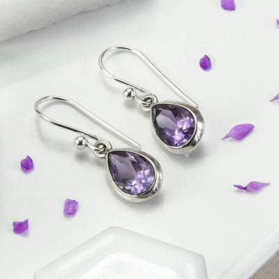White metal purple teardrop bezel hook earrings on white tile with petals