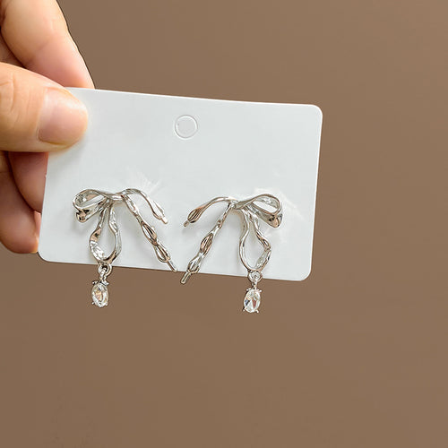 Silver liquid bow clear drop earrings on white card against brown wall