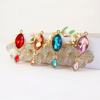 Four fleur-de-lis ornaments with red, peach, blue, and pink faceted stones stand against a white display.