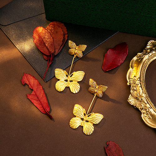 Gold butterfly dangle earrings on brown surface with dried red leaves