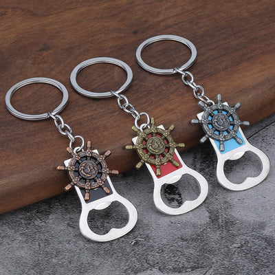 Three ship wheel bottle opener keychains in copper, bronze, and silver tones rest on a wood surface.