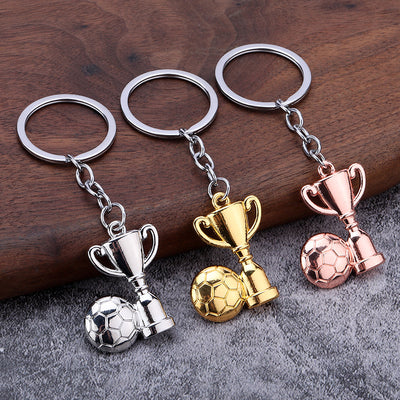 Three cup and soccer ball keychains in silver, gold, and rose gold on a wood and gray surface.