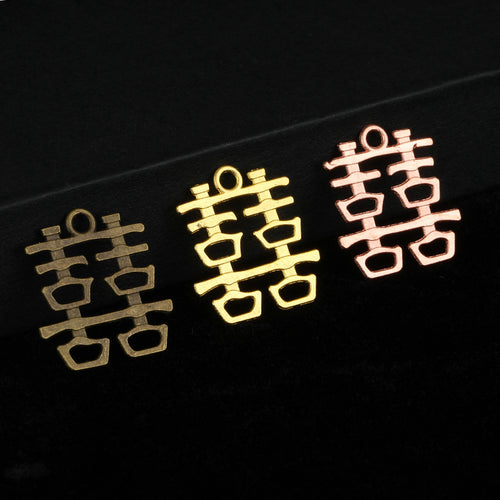 Three double happiness charms in antique bronze, gold-tone and rose gold-tone side by side on a black box