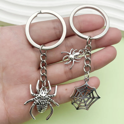 Two silver-tone keychains rest on a hand, one with a large spider and one with a spider web plus small spider.