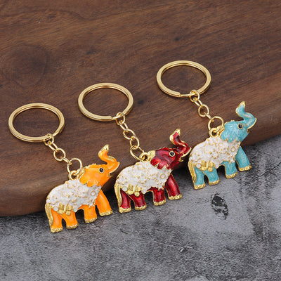 Three gold-tone elephant keychains in orange, red, and light blue on a wooden surface.