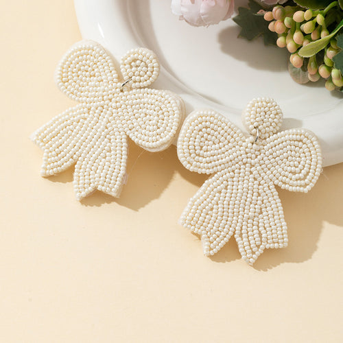 White bow beaded drop earrings shown on cream background with pink flowers nearby