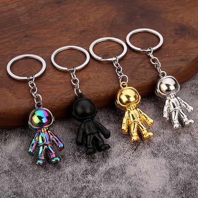 Four astronaut figure keychains in rainbow, matte black, gold, and silver finishes on wood and gray fabric.