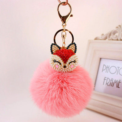 A red rhinestone fox face sits above a coral pink fluffy pom pom and gold-tone key hardware.
