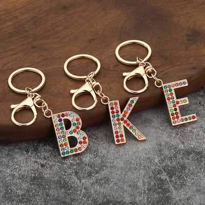Three gold-tone keychains display the rhinestone letters B, K, and E on a wooden surface.