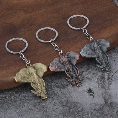 Three elephant-shaped keychains in brass, copper, and silver tones displayed on a wooden surface.