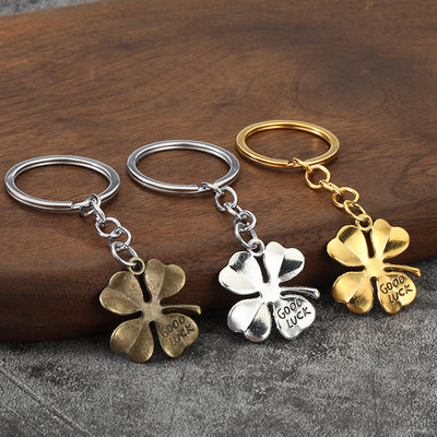 Three four-leaf clover keychains in antique bronze, silver, and gold tones on a wood surface.