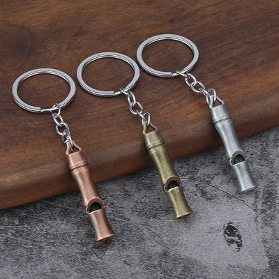 Three metal whistle keychains in antique copper, bronze, and silver tones on a wood surface.