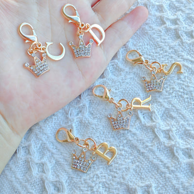 Overview of four gold-tone crown keychain charms with different rhinestone crowns and letter charms held in a hand