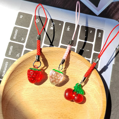 Three resin fruit phone straps: red strawberry, clear strawberry, and red cherries on wooden dish