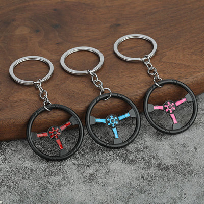 Red, light blue, and pink steering wheel keychains arranged on a wood and gray surface.