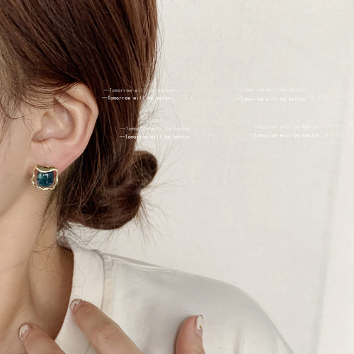 Model wearing blue-green square stud with gold wavy rim