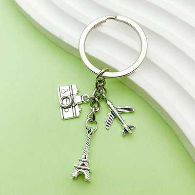 A silver-tone keychain with dangling Eiffel Tower, airplane, and camera charms on a green background.