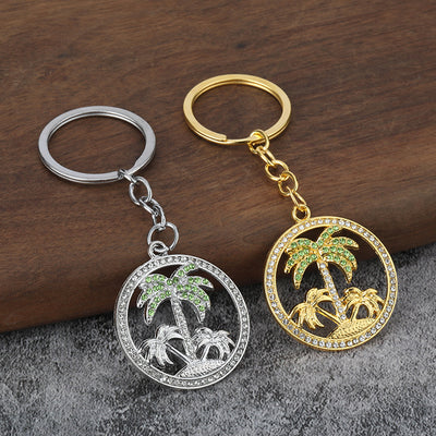 Gold-tone and silver-tone palm tree keychains rest side by side on a wooden and gray fabric surface.